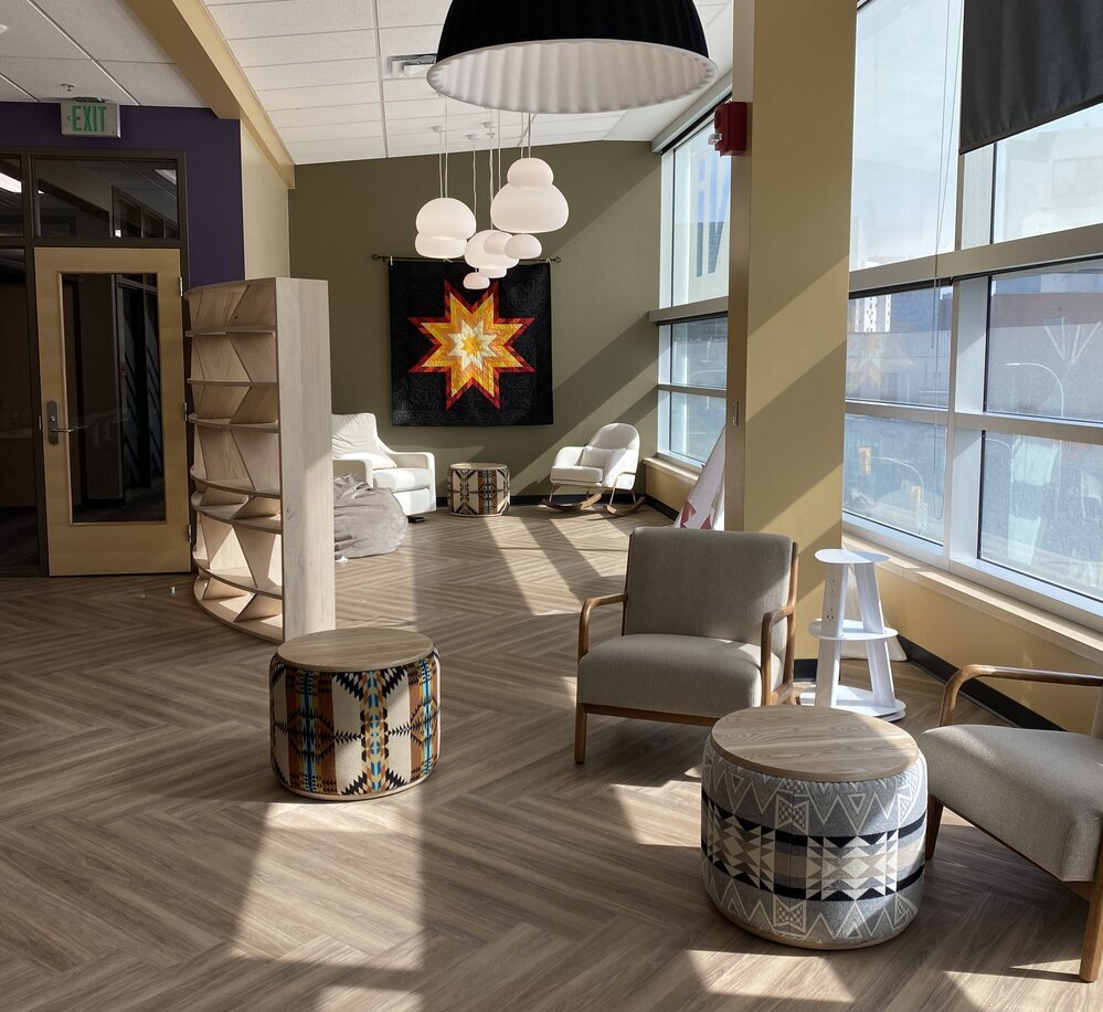 Detail photo of a corner of Aabijijiwan New Media Lab. Natural light pour into the room from a wall of windows to the right. On the far wall is a star blanket tapestry and a custom bookshelf. In the foreground of the photo are custom wood upholstered chairs and drum stools upholstered with fabric inspired by Anishinaabe geometric patterns.