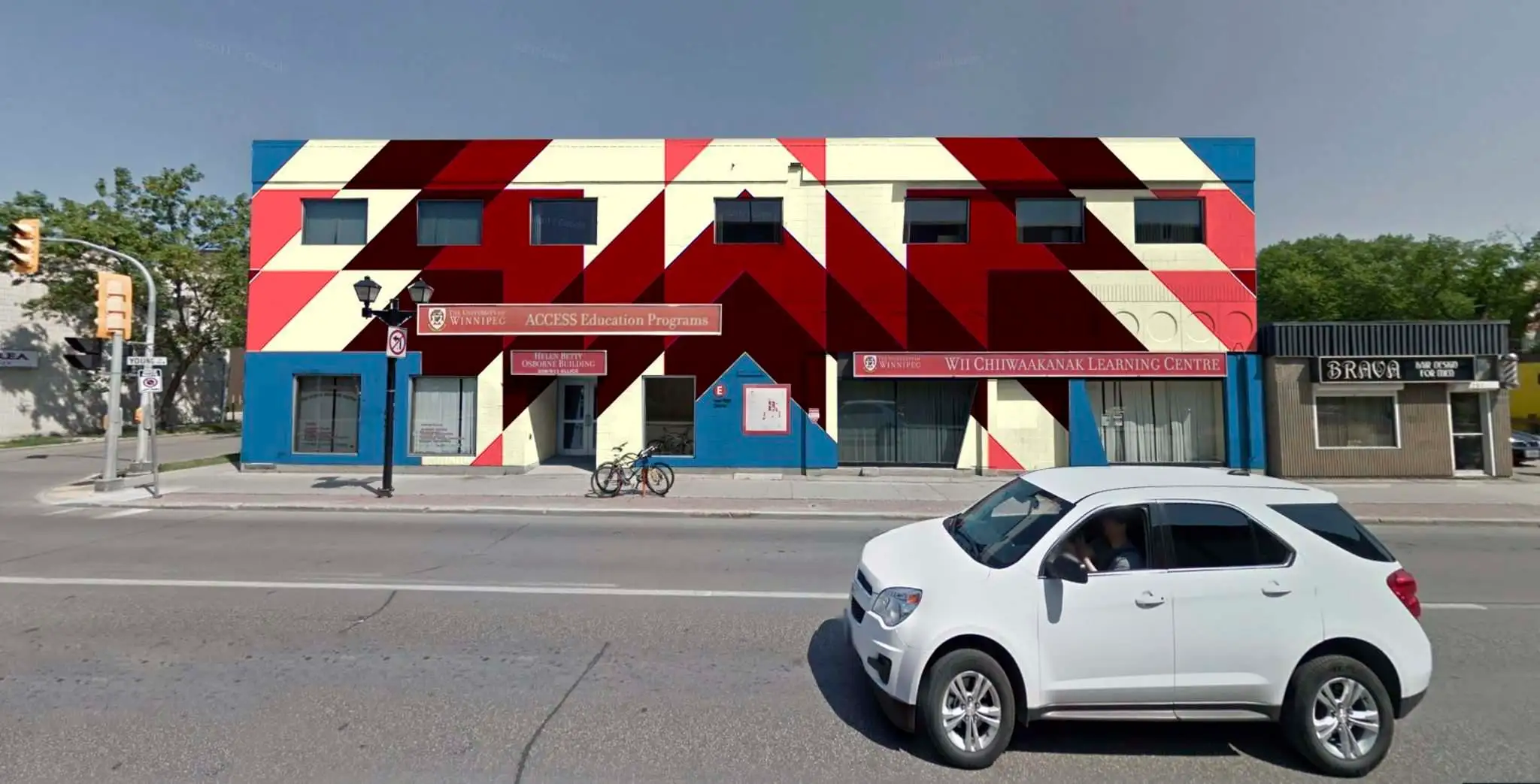 Exterior elevation of the Helen Betty Osborne Building at 511 Ellice Avenue. The building is cloaked in a brightly coloured geometric mural of a star blanket in sky blue, bright red, deep red, maroon, and white.