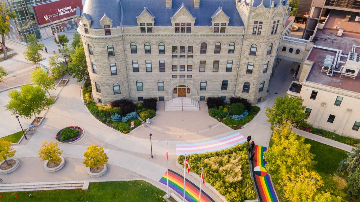 Aerial view of Portage Commons at University of Winnipeg. There is a clear view of the front facade of Wesley Hall, a castle-like building. On the pavement is a sidewalk mural of three pride flags.