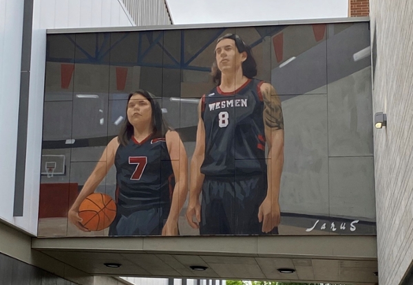 Photo of mural by Emmanuel Jarus on the wall connecting Duckworth Centre and the Axworthy Heath and RecPlex at the University of Winnipeg. The mural depicts two Indigneous basketball players in Wesmen uniforms, they stand in a gymnasium. A shorter woman stands to the left of a tall man with a sleeve tattoo.