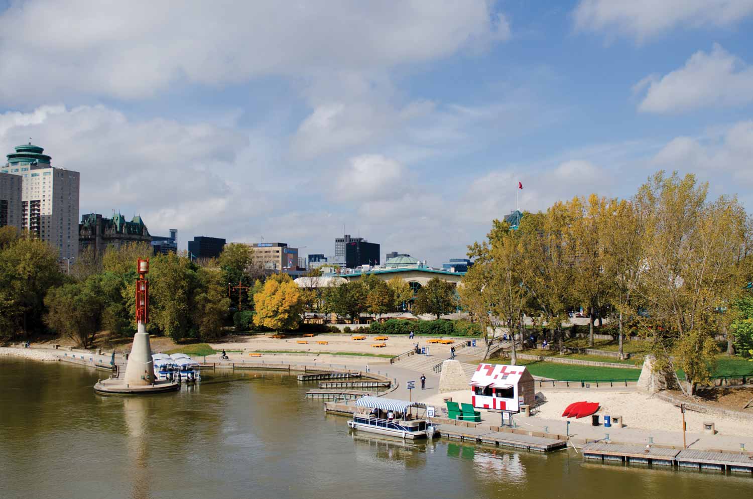 A view from the water at the Forks Historic Port. A few boats are on the water. Yellow benches are scattered along the dock. The Forks Market is in the back.