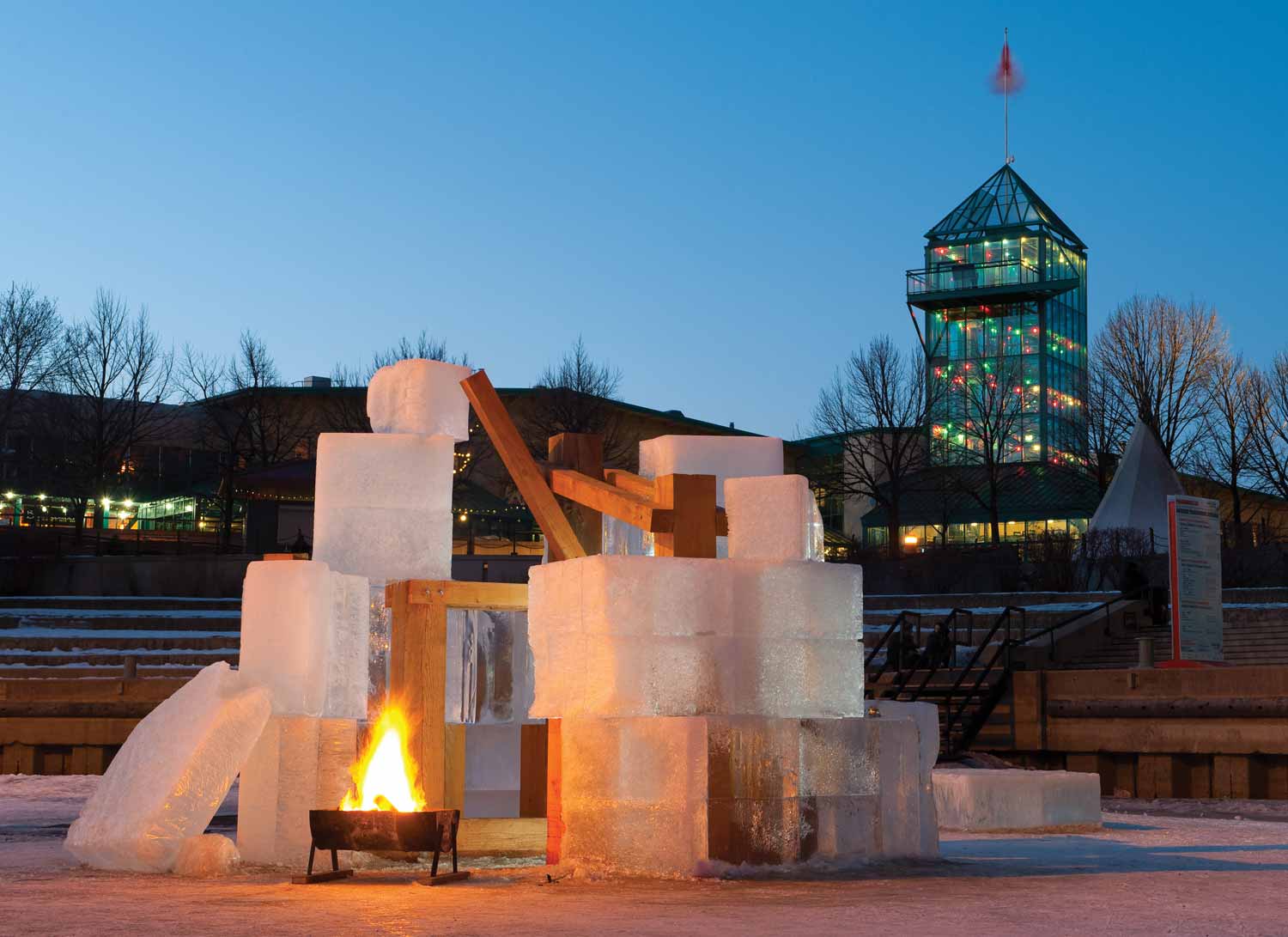 At dusk, a fire burns in a fire pit next to a structure built of large cubes of ice and blocks of wood. The Forks Market Tower is in the background.
