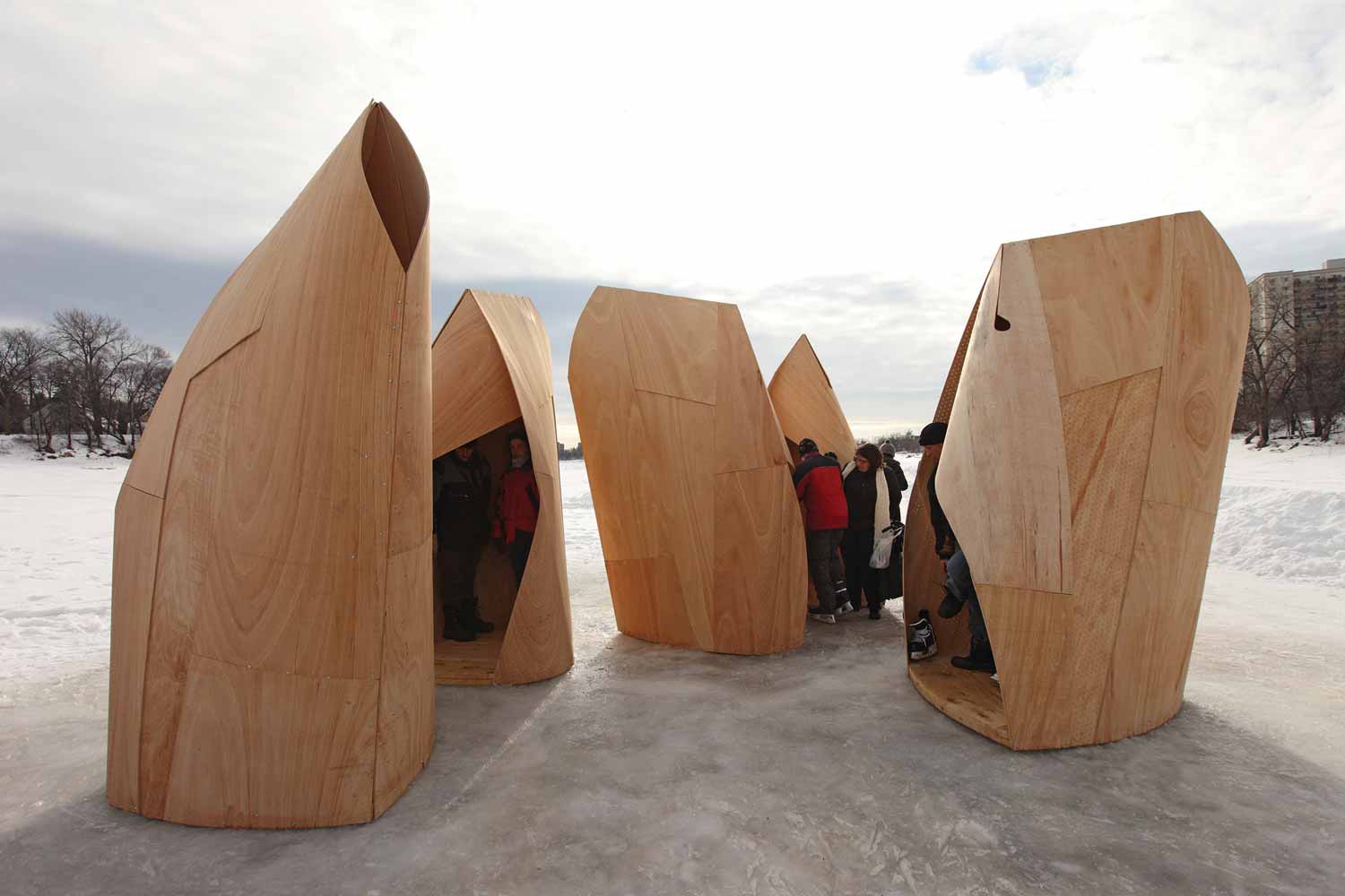 Multiple uniquely shaped huts made of flexible moulded plywood are clustered on the river trail. Skaters are visible inside some, and are entering the openings in others.