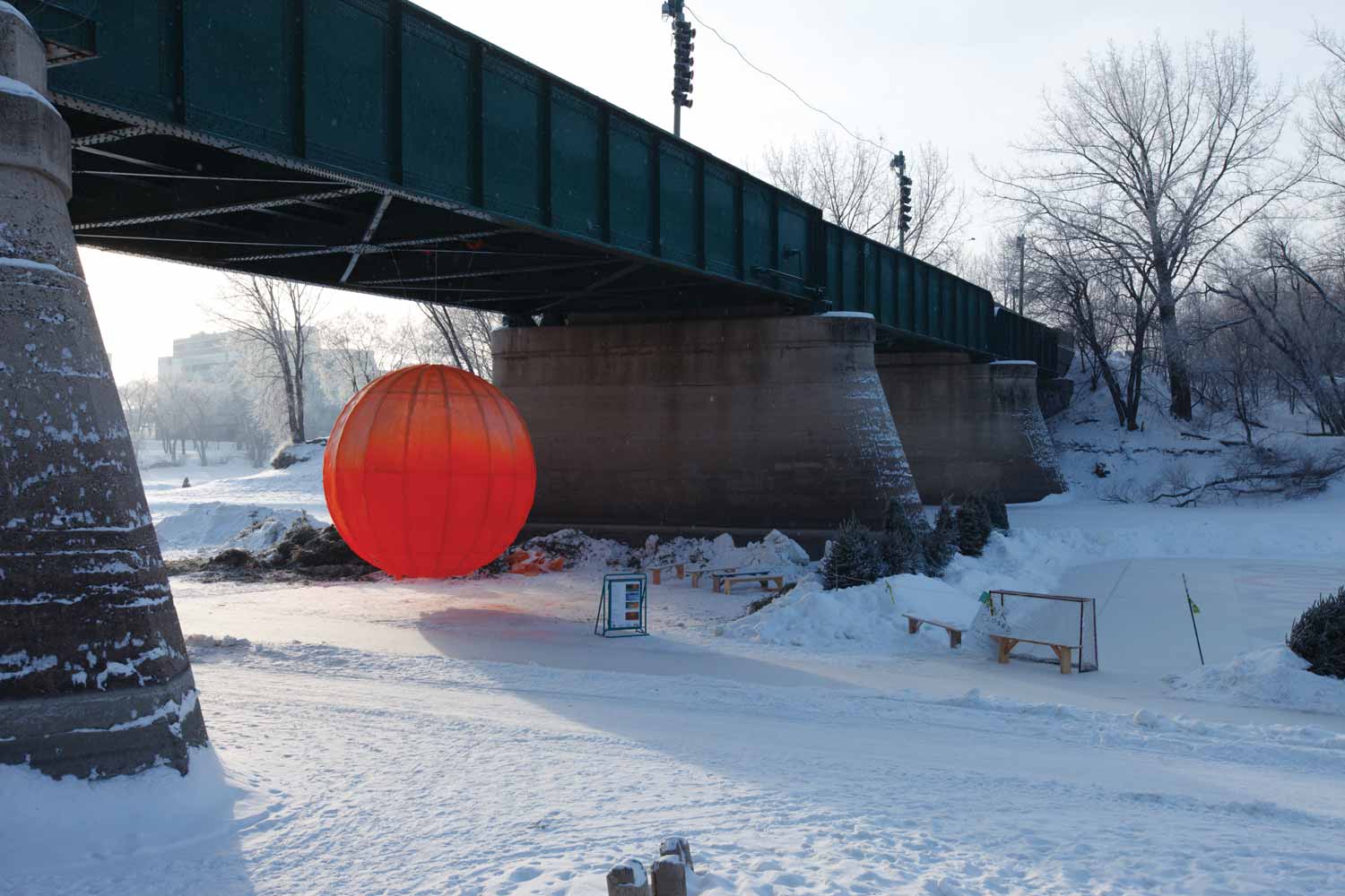 A large orb made of red fabric on a metal frame hangs underneath The Forks Historic Rail Bridge over the skating trail.
