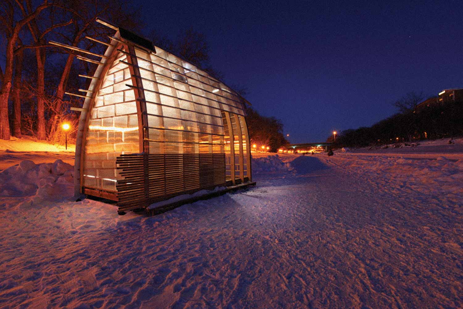 A hut with wooden frame, polycarbonate and aluminum panels, is lit up from the inside on the dark river trail.
