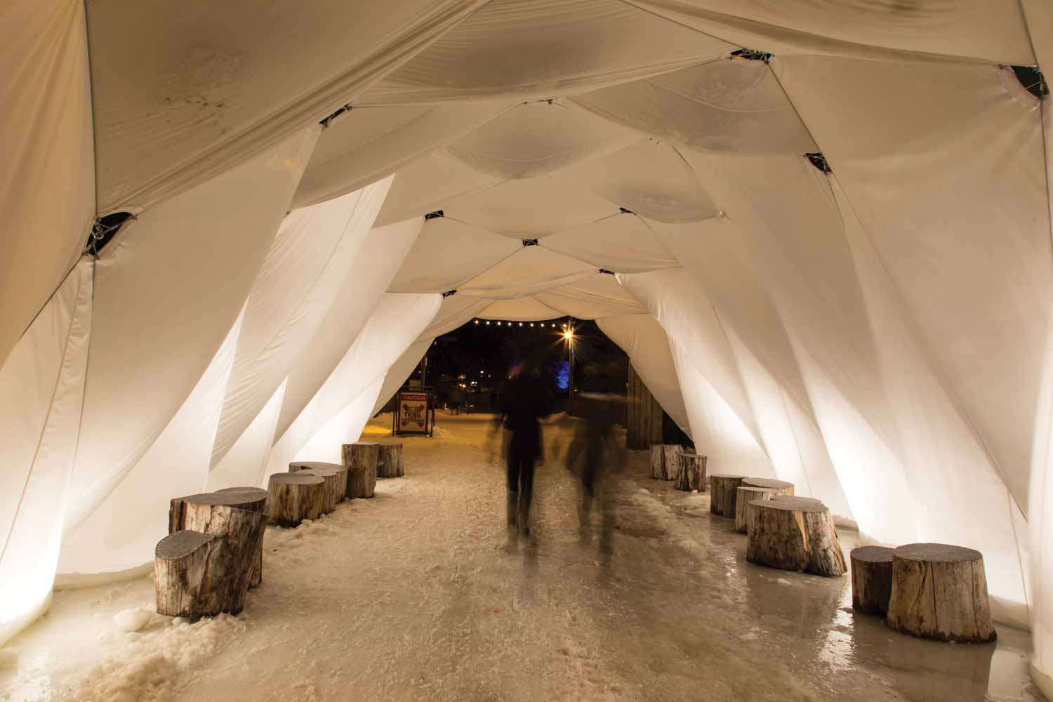 Two people skate along the river trail through a white tent that looks folded like origami. Stumps of wood line the trail.
