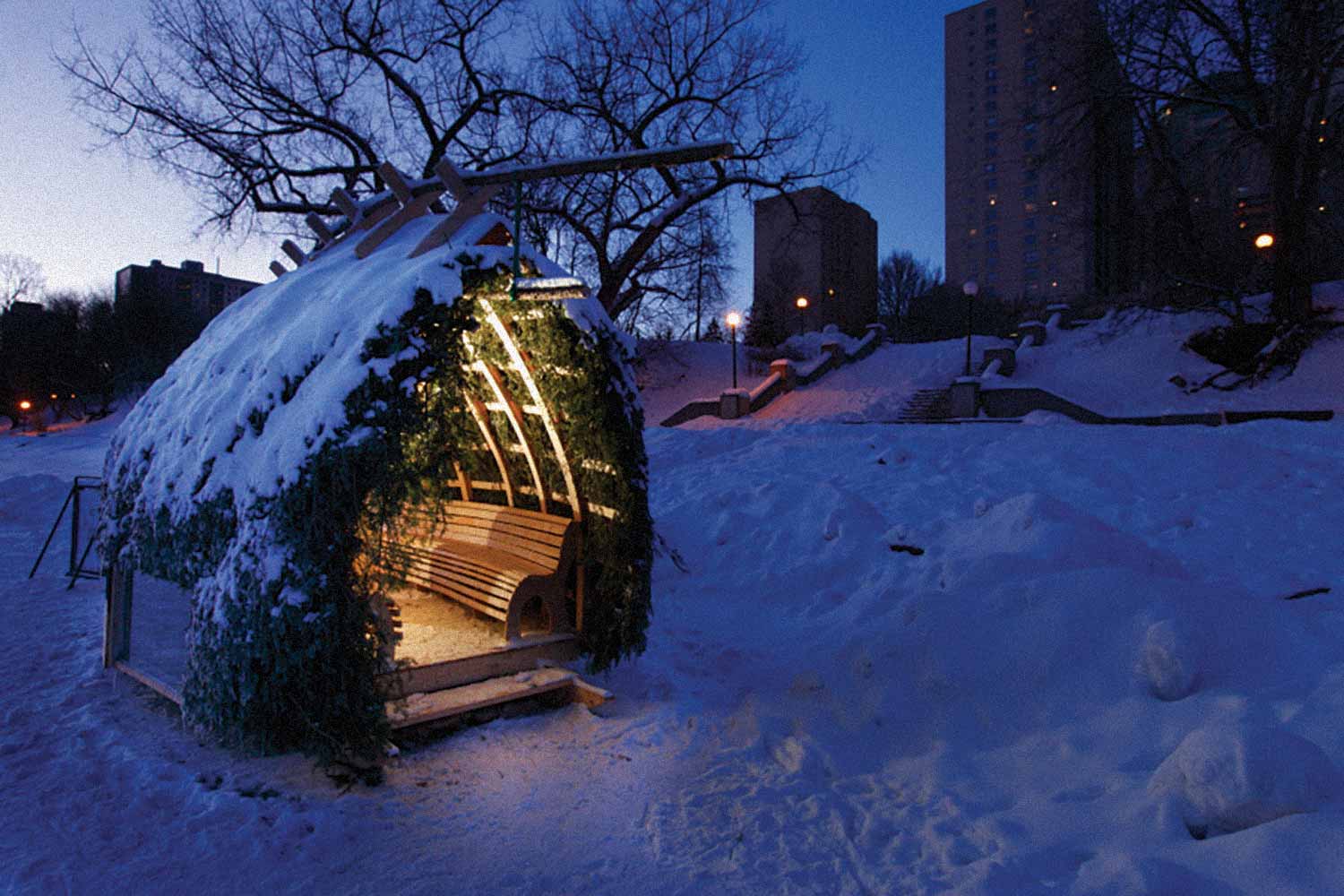 A small hut made of wood is lit up at night on the river trail. Its roof is covered in fir tree branches.