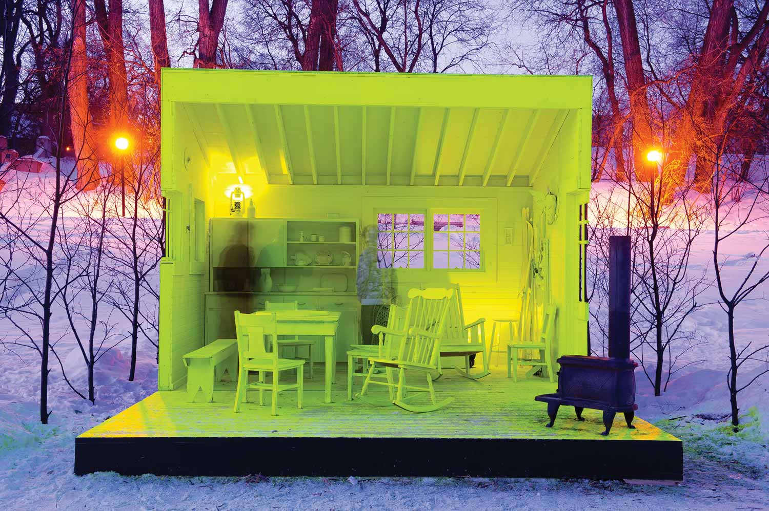 A cabin room cut in half on a base on the river. A table and chairs and other room decorations are all painted fluorescent yellow. A wood stove sits in the corner. A skater from the river trail is standing next to a bench in the warming hut