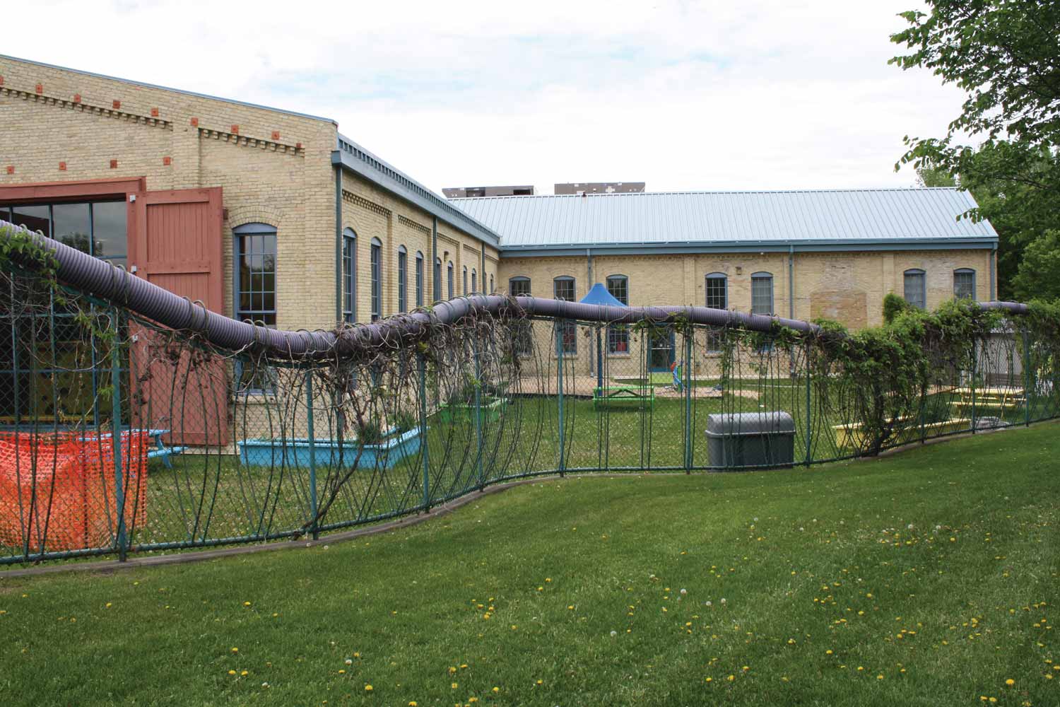 The Children's Museum in 2015, shown from the south-southeast. A wavy fence around the building contains a grassy area with garden boxes and a picnic table.