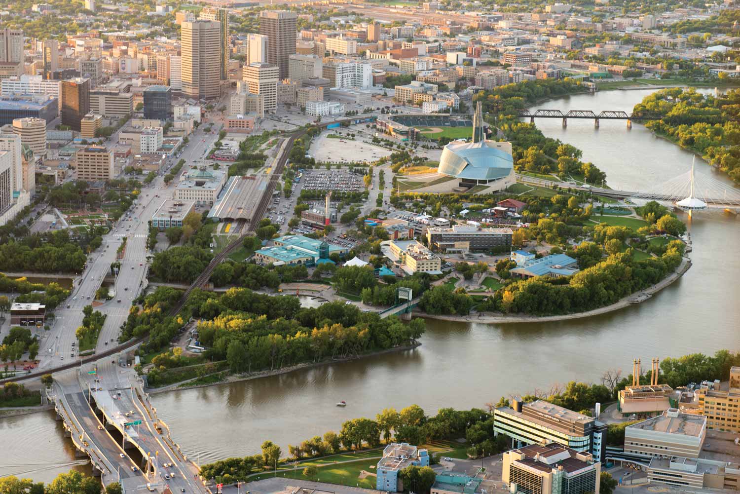 Aerial photo of the Forks at dusk in the summer, with Main Street on the left and the Red River on the right. Various landmarks are pictured, including the Canadian Museum for Human Rights.