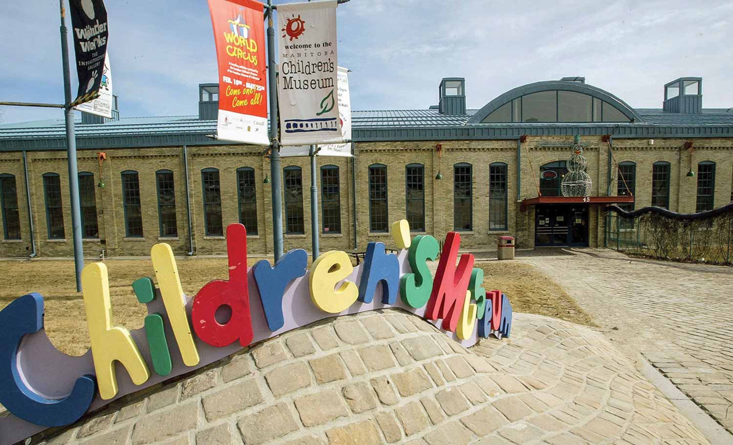 The Manitoba Children's museum in 1994, showing the original brick engine house with a blue metal roof. There is a wavy path out front with a colourful lettered sign reading "Children's Museum"