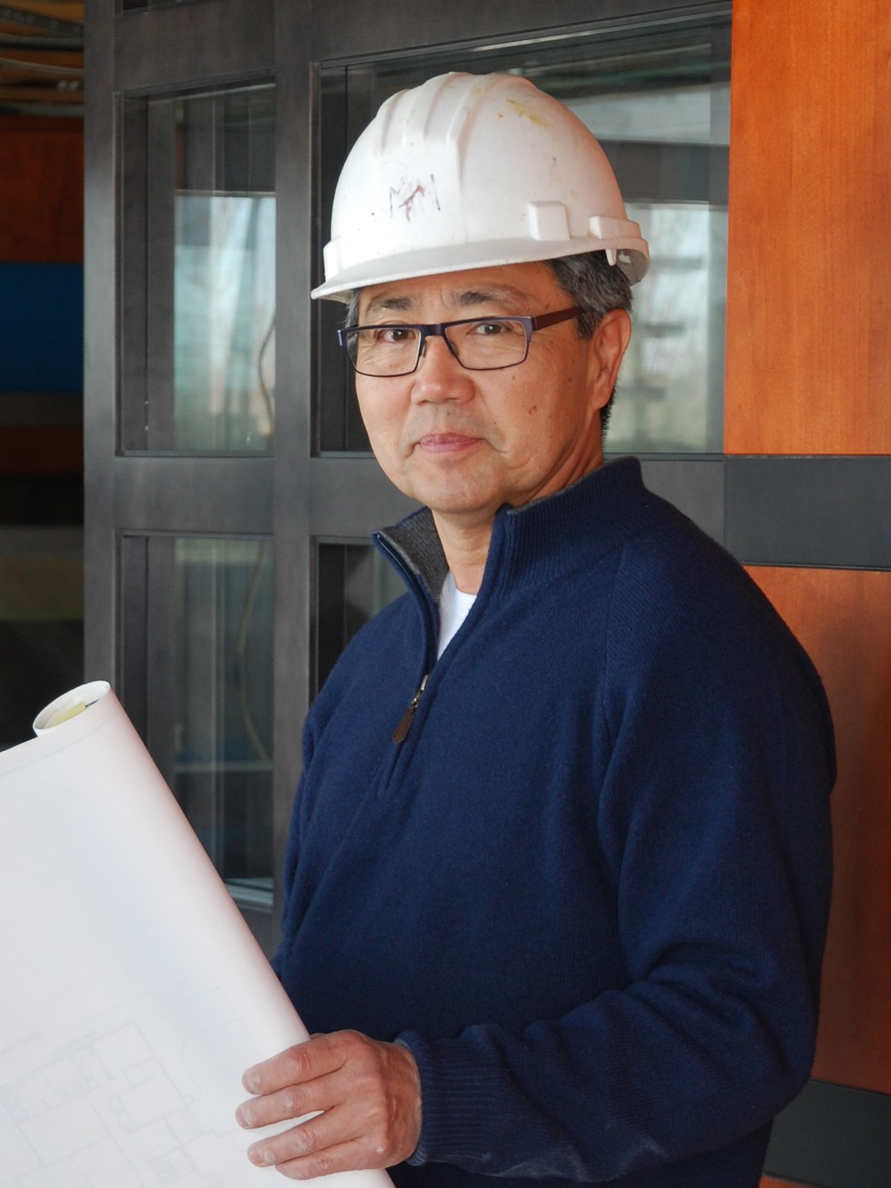 Image shows a man with glasses and a white hard hat. He is wearing a blue sweater and holding a large roll of paper.