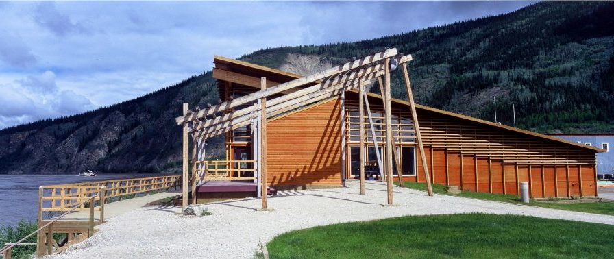 Image shows a contemporary structure with bright amber wood panelling and a sharp, ascending roofline. A wooden sculpture sits in front of the structure.
