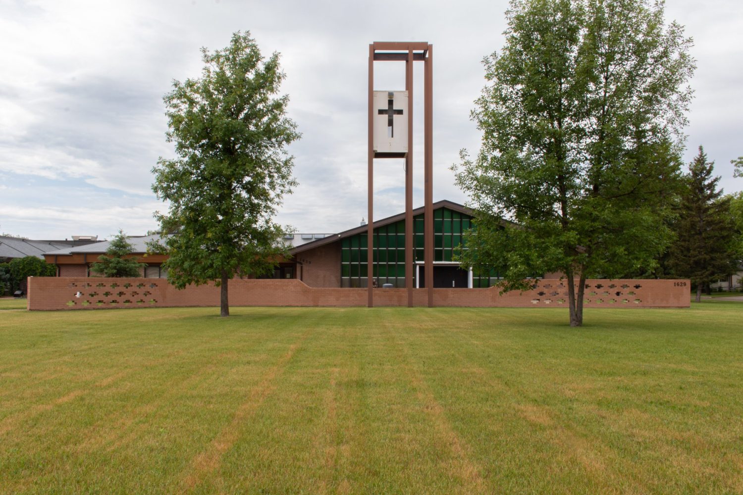 Image shows the exterior of St. Vital Church. A long and brown brick fence spans across the lot, punctuated with cross shaped indentations on the far left and right sides. The building has a dramatic and sloping triangular frame with large glass windows. A cross is carved onto a white and rectangular box, which is suspended on brown beams.