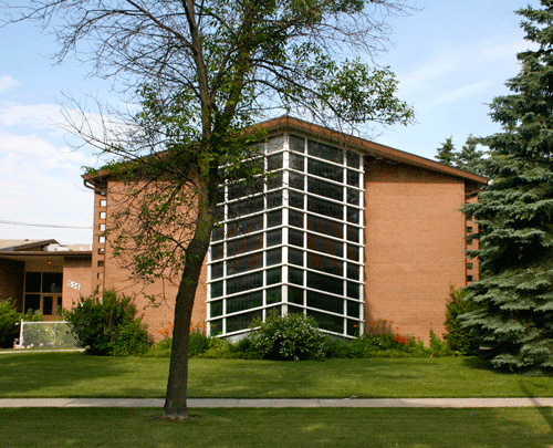 Image shows the brown brick exterior of St. Joseph the Worker Church. It has a large window at the centre and a series of white and triangular grilles.