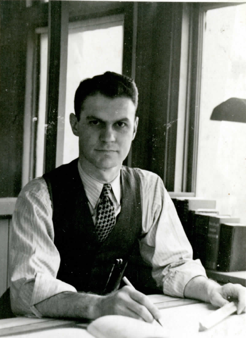 Image shows a man with a vest and a tie. He is sitting at a desk, holding a pencil and smiling slightly at the camera.
