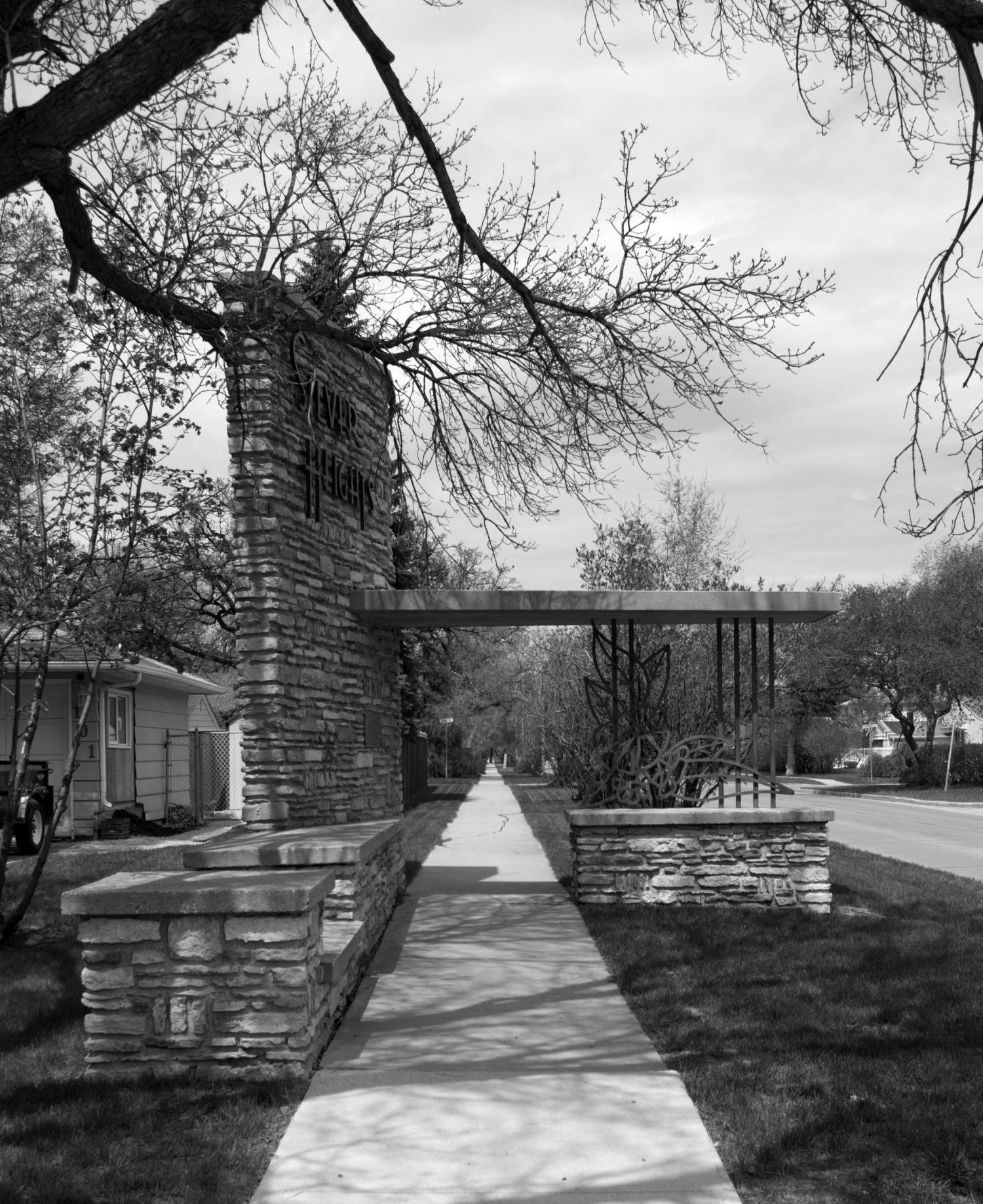 Image shows a black and white image of Silver Heights Gates from the sidewalk. It prominently shows a iron wrought flowers.