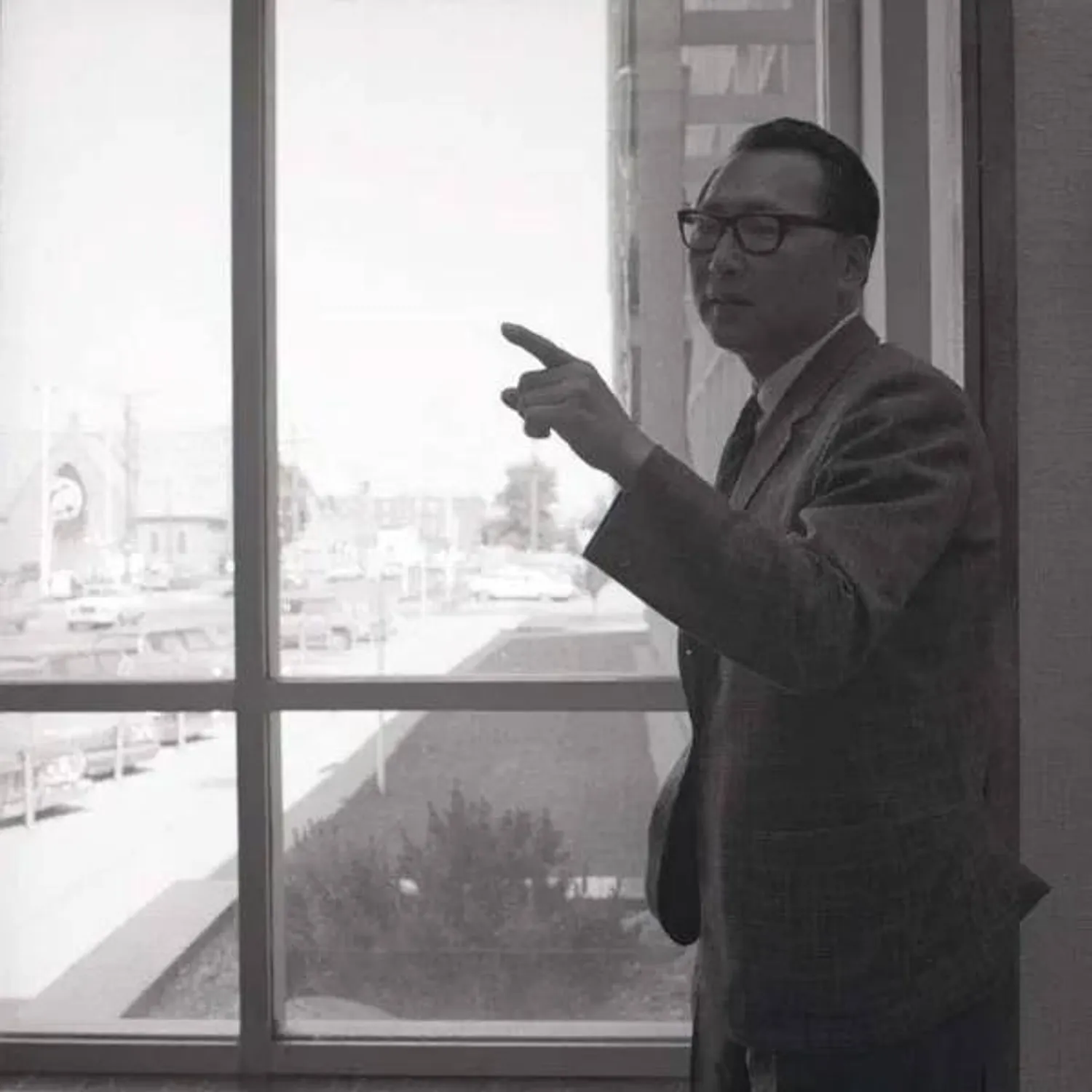 A man with glasses and a suit stands in front of a window. He is pointing at something beyond the image.