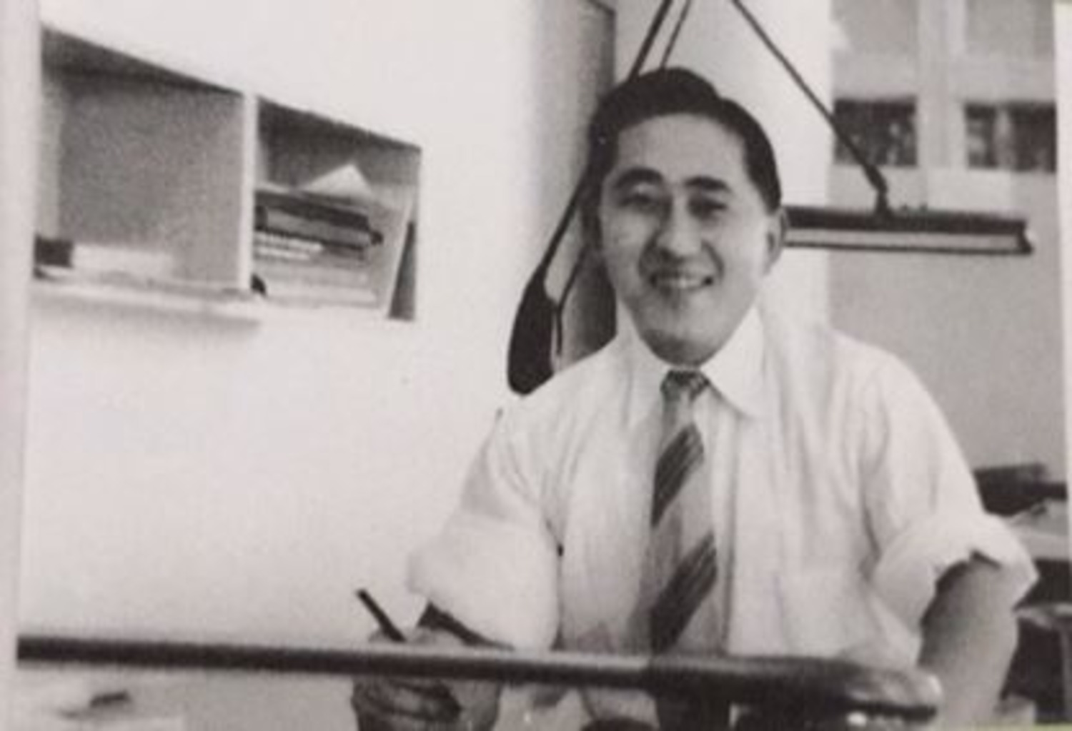 Image shows a smiling man with a tie, holding a pencil over a desk.