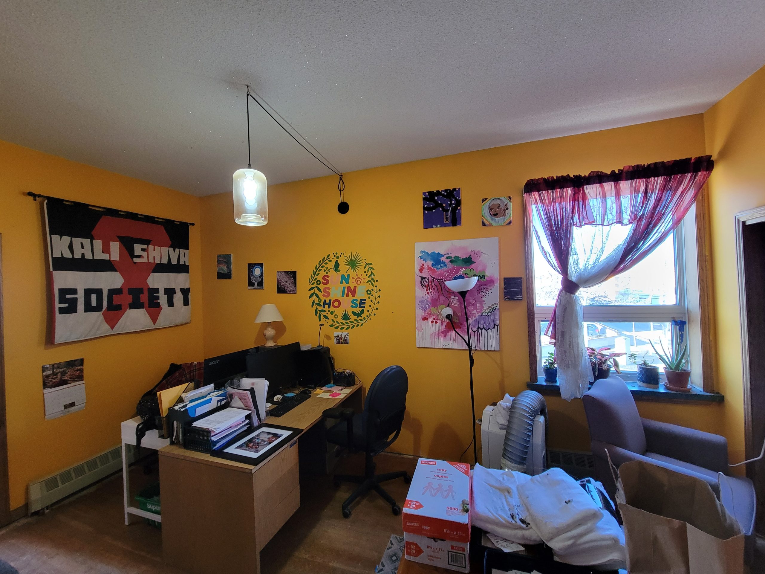 Office space on the upper floor of Sunshine House. The walls are painted bright yellow and there is a black, white, and red flag on the wall that says "Kali Shiva Society".