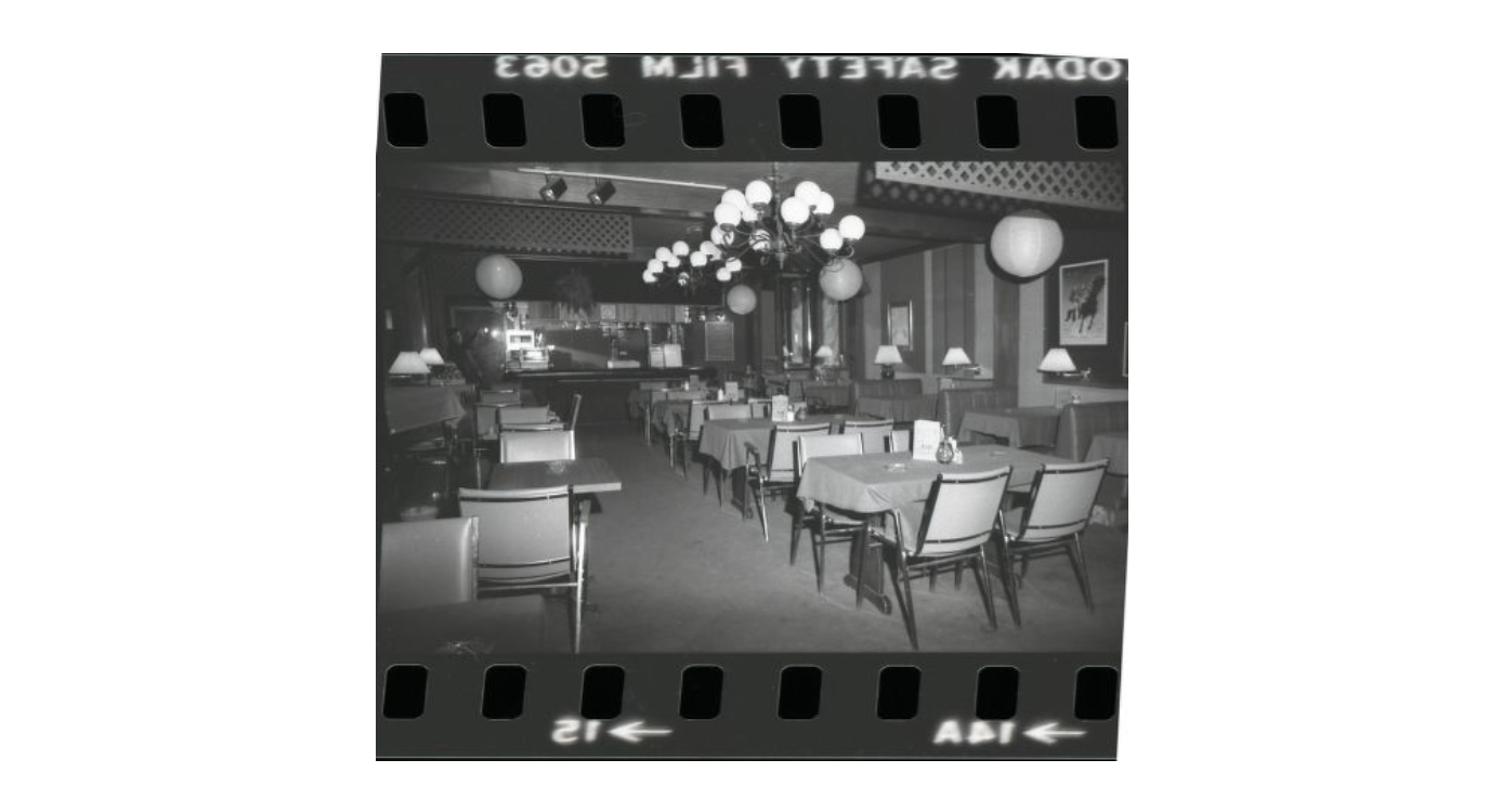 A black and white negative of the interior of Giovanni's Room. There are rows of smalls tables and a few large light fixtures can be seen hanging from the low ceiling.