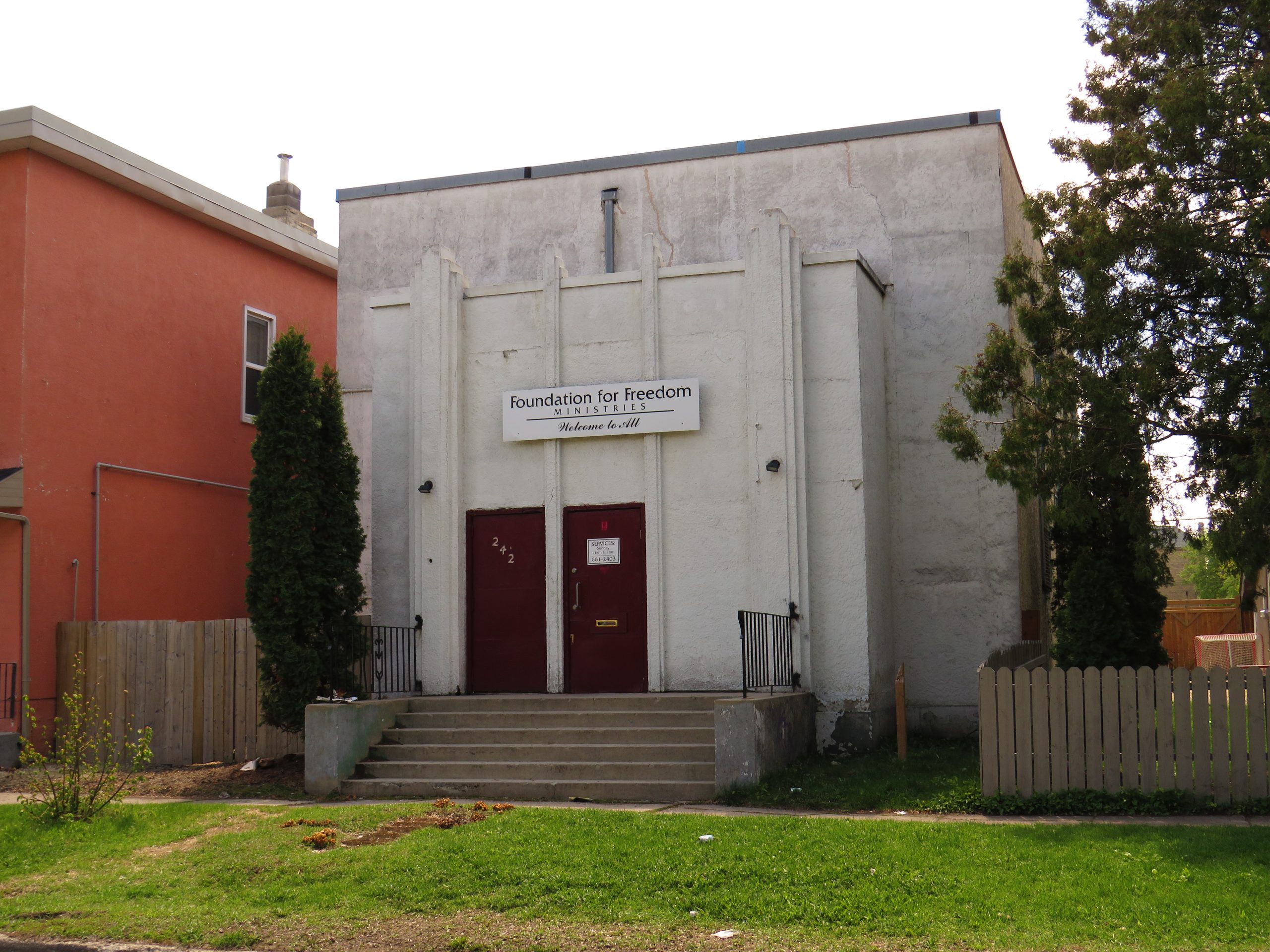 The exterior of 242 Manitoba Ave. The building is light grey stucco with no windows, and the two front doors are red. There is a sign above the doors that says "Foundation for Freedom Ministries - Welcome to All".