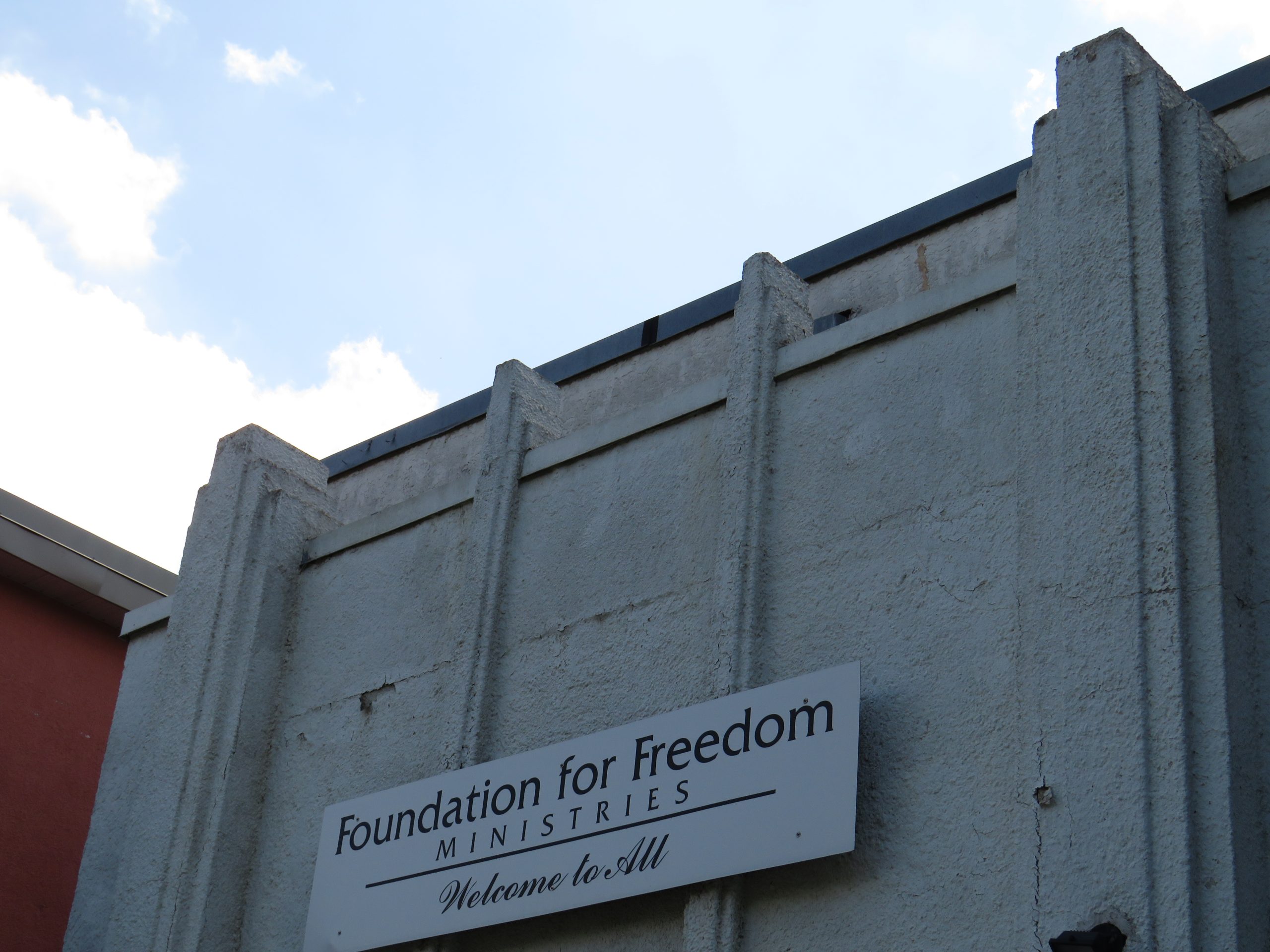 A closeup of the exterior of 242 Manitoba Ave. The building is light grey stucco with no windows. There is a sign above the doors that says "Foundation for Freedom Ministries - Welcome to All".