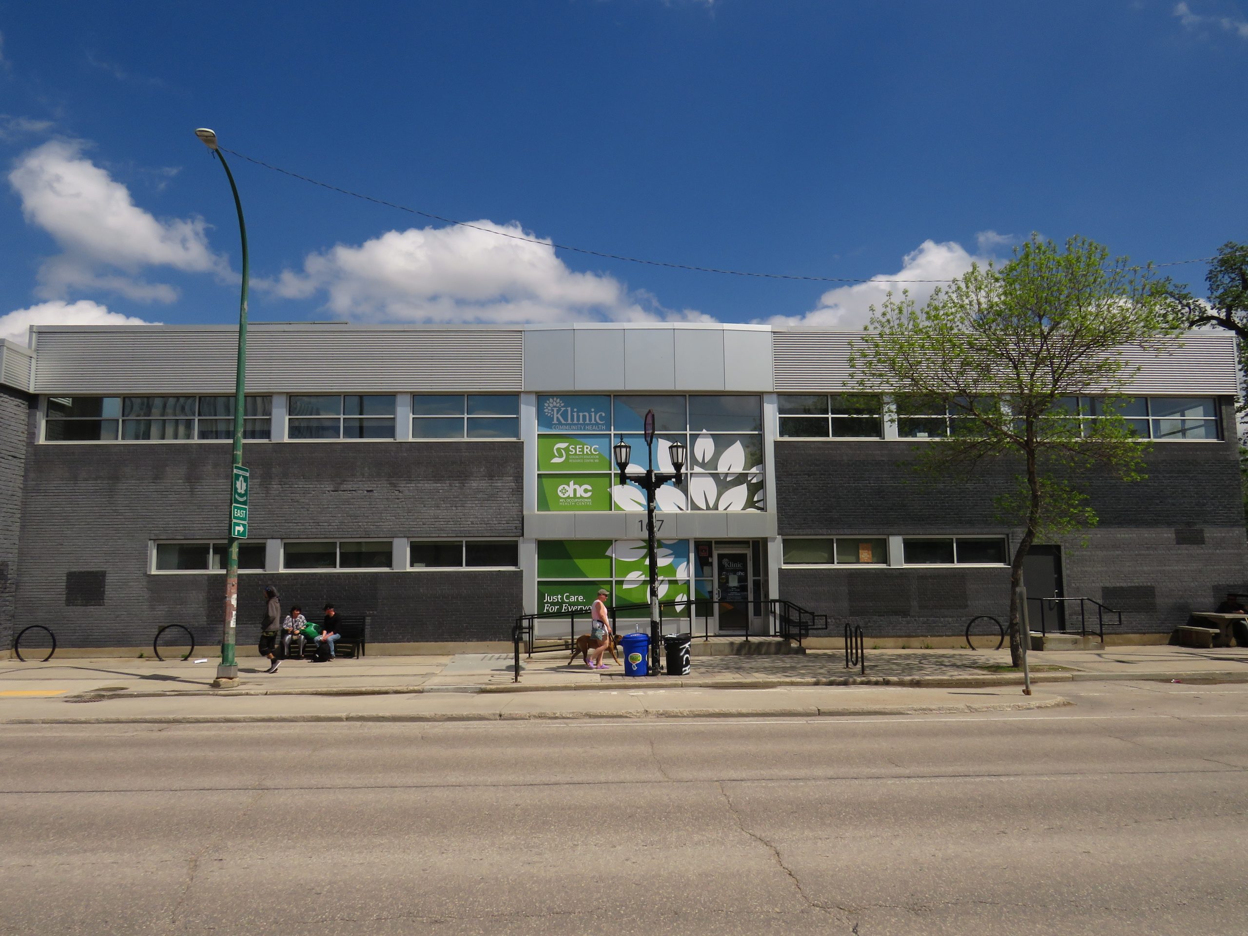 The exterior of 167 Sherbrook St. The building is grey brick with a large green and blue sign in the window listing the organisations in the building, Klinic, SERC, and OHC.
