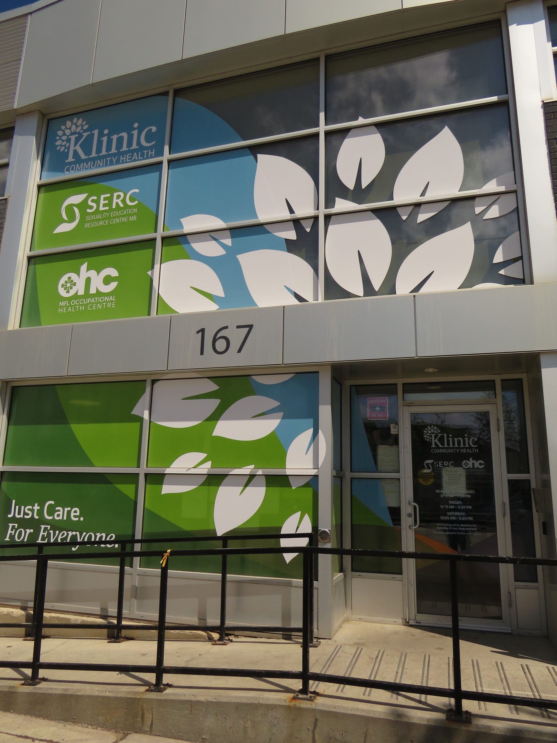 A closeup of the exterior of 167 Sherbrook St. There is a large green and blue sign in the window listing the organisations in the building, Klinic, SERC, and OHC.