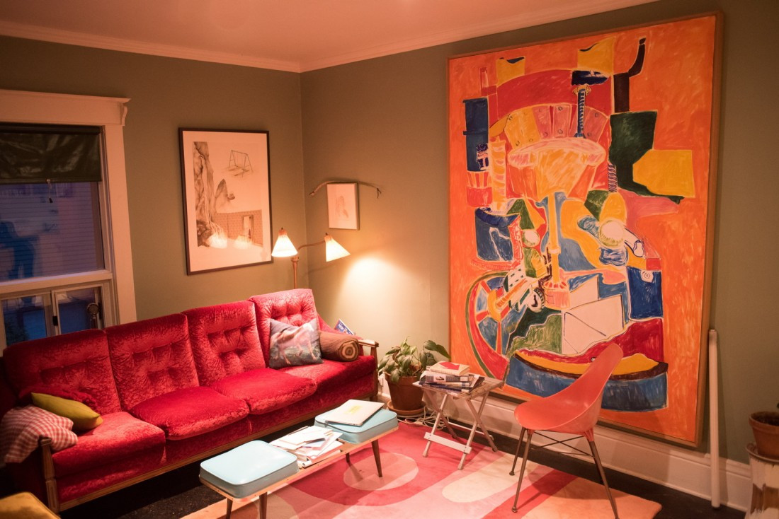 The interior of Homo Heaven. There is a red couch and a pink and red rug on the floor, and a large, bright coloured painting hangs on the greenish-beige wall.