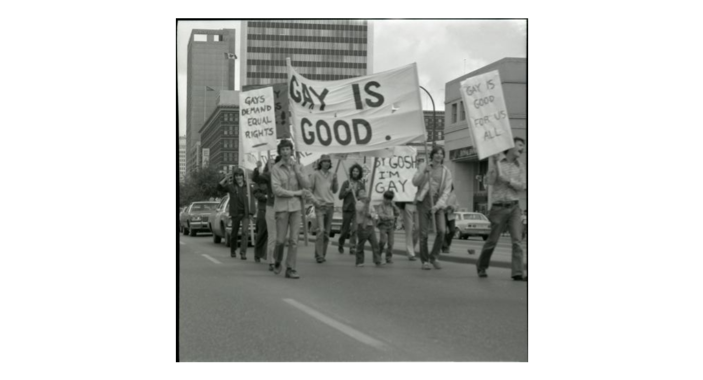 A black and white negative of Gay Pride March through Downtown Winnipeg. Demonstrators can be seen holding various signs with different slogans including two men carrying a large banner reading "Gay is Good".